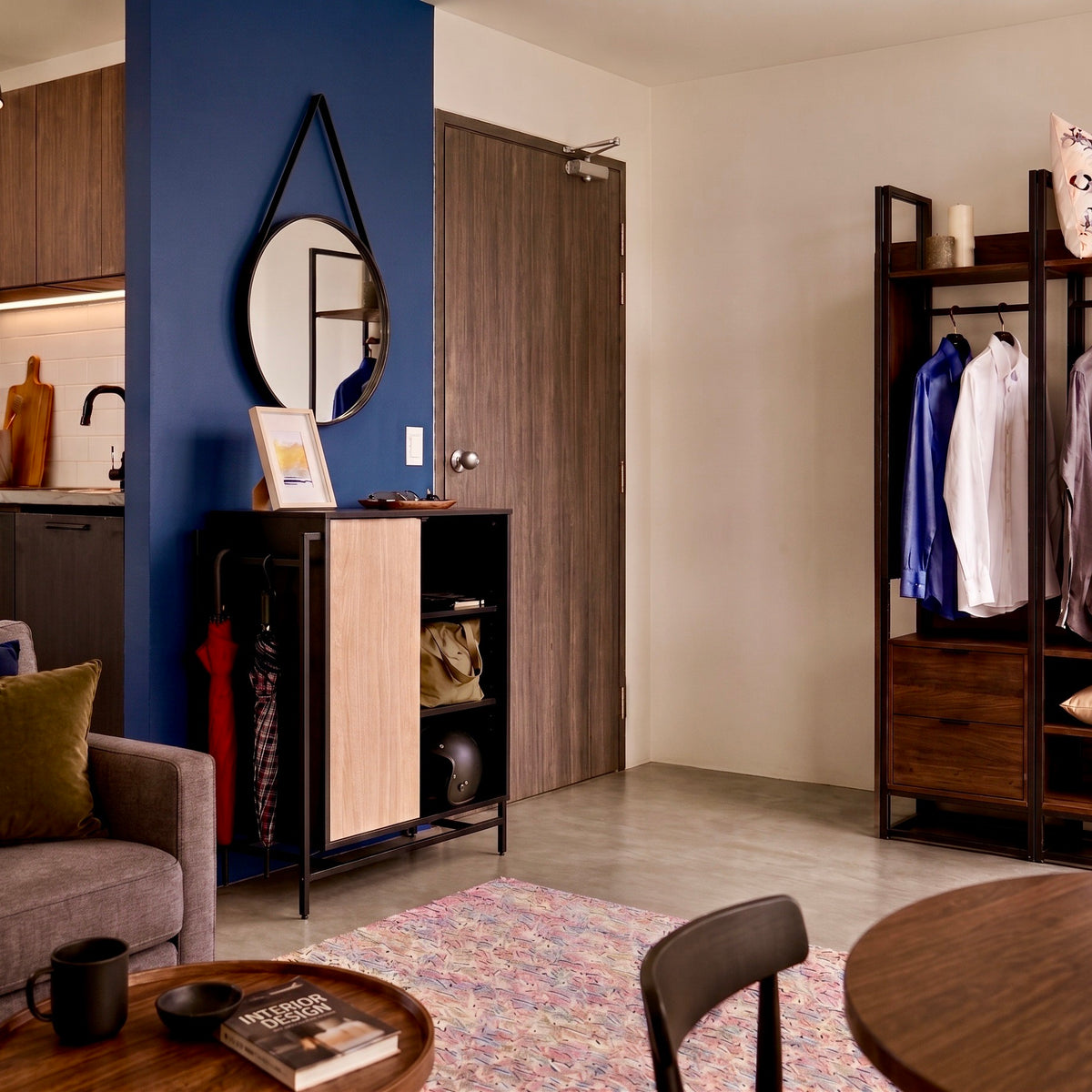 A modern, stylishly decorated apartment entryway featuring a dark wood door, a blue accent wall with a black-framed mirror, a black and light wood entry cabinet, and a clothing rack with blue and white shirts.