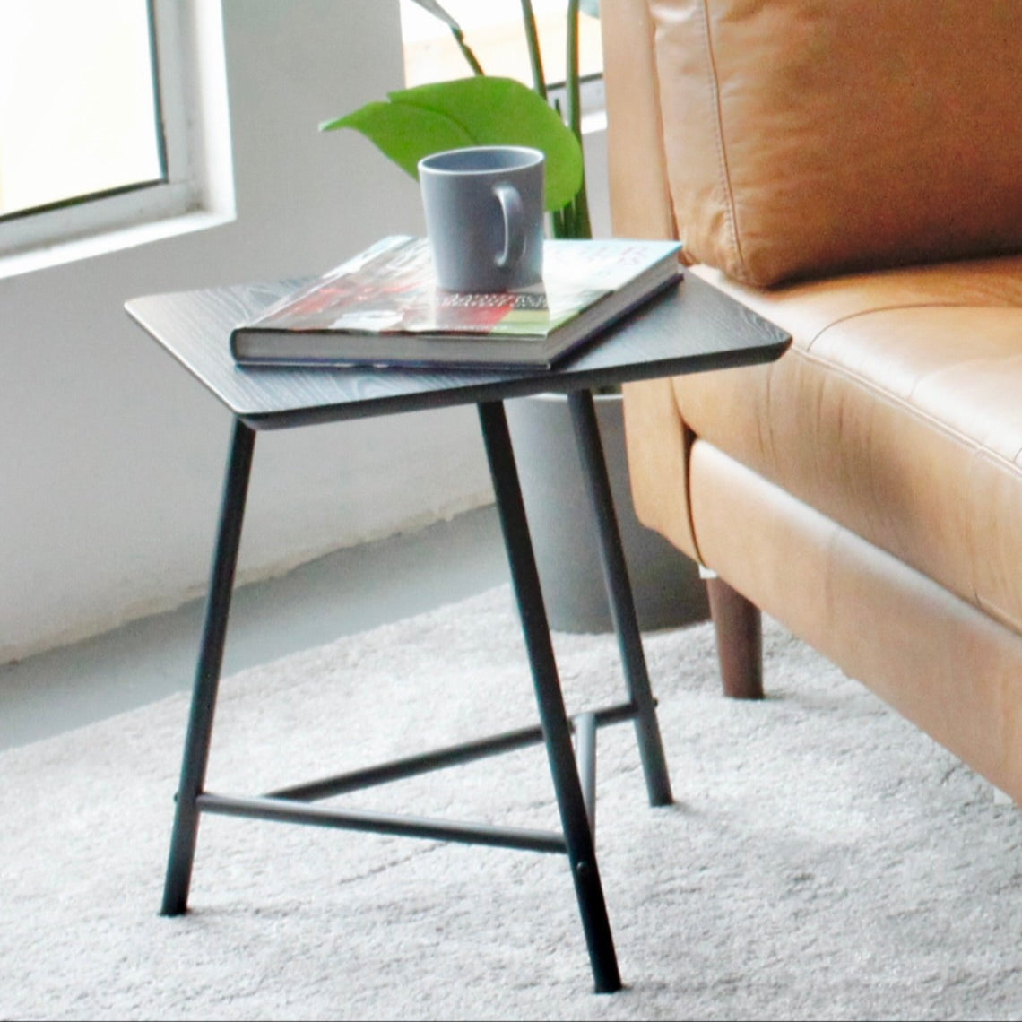 Beige leather sofa with a small black table holding a book and a mug, in a bright room with large windows.