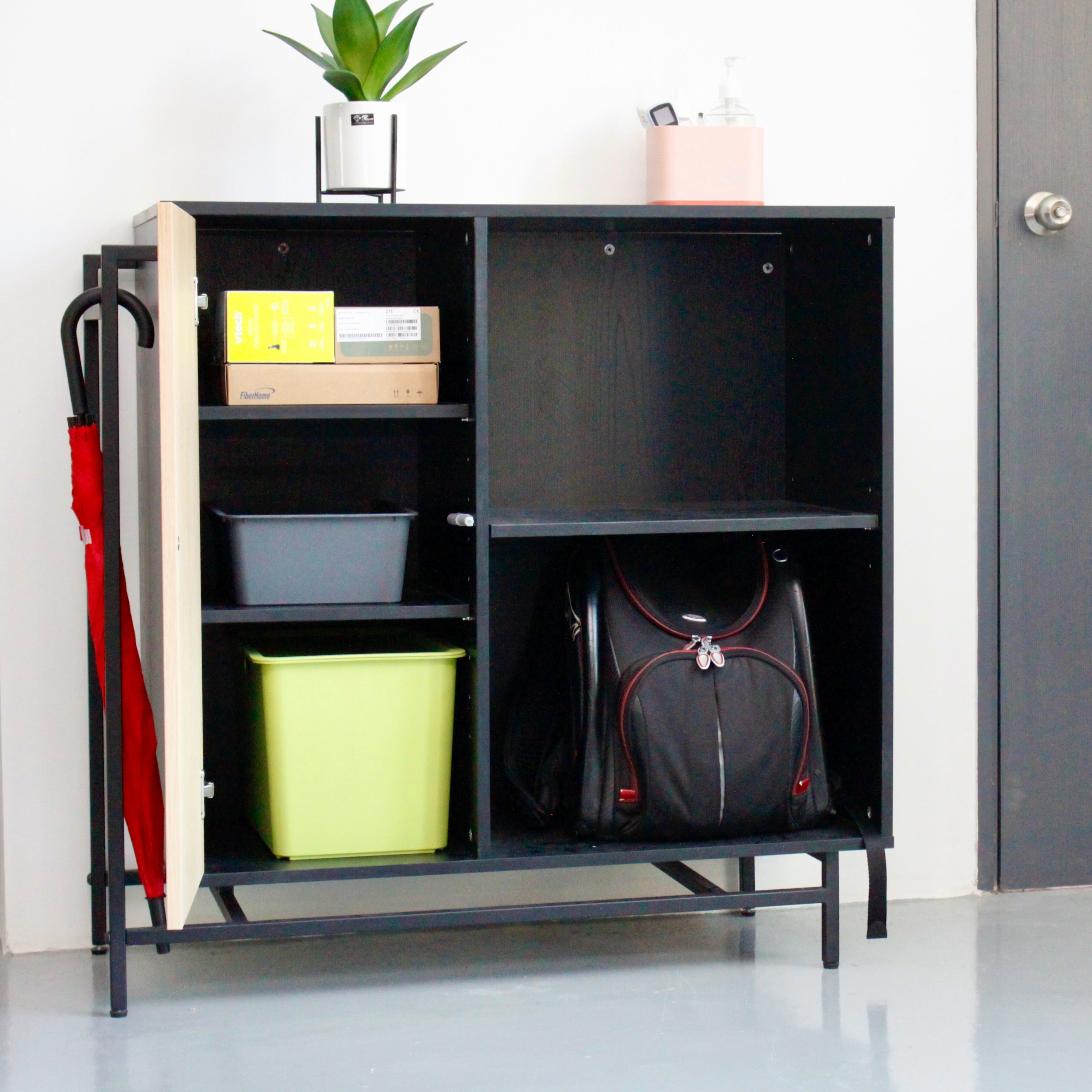 A black storage cabinet with a metal frame, featuring a natural oak door and multiple compartments with items such as a green bin, a black bag, and a red umbrella stored inside.