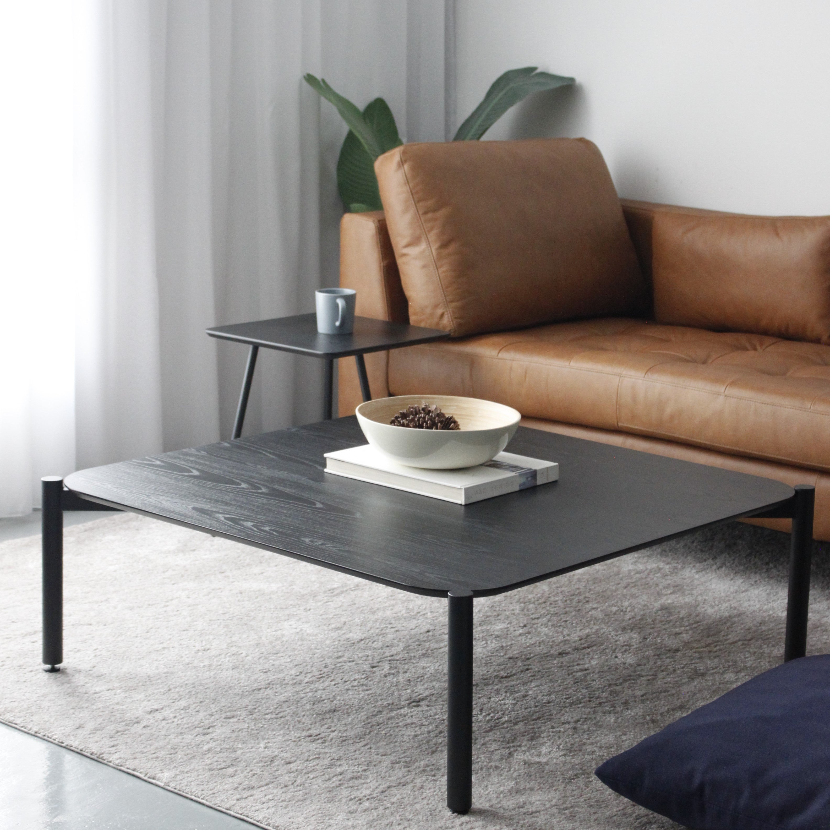A black square coffee table with a wood grain effect top, placed on a grey area rug next to a brown sofa, with a plant and a cup on top of the table.