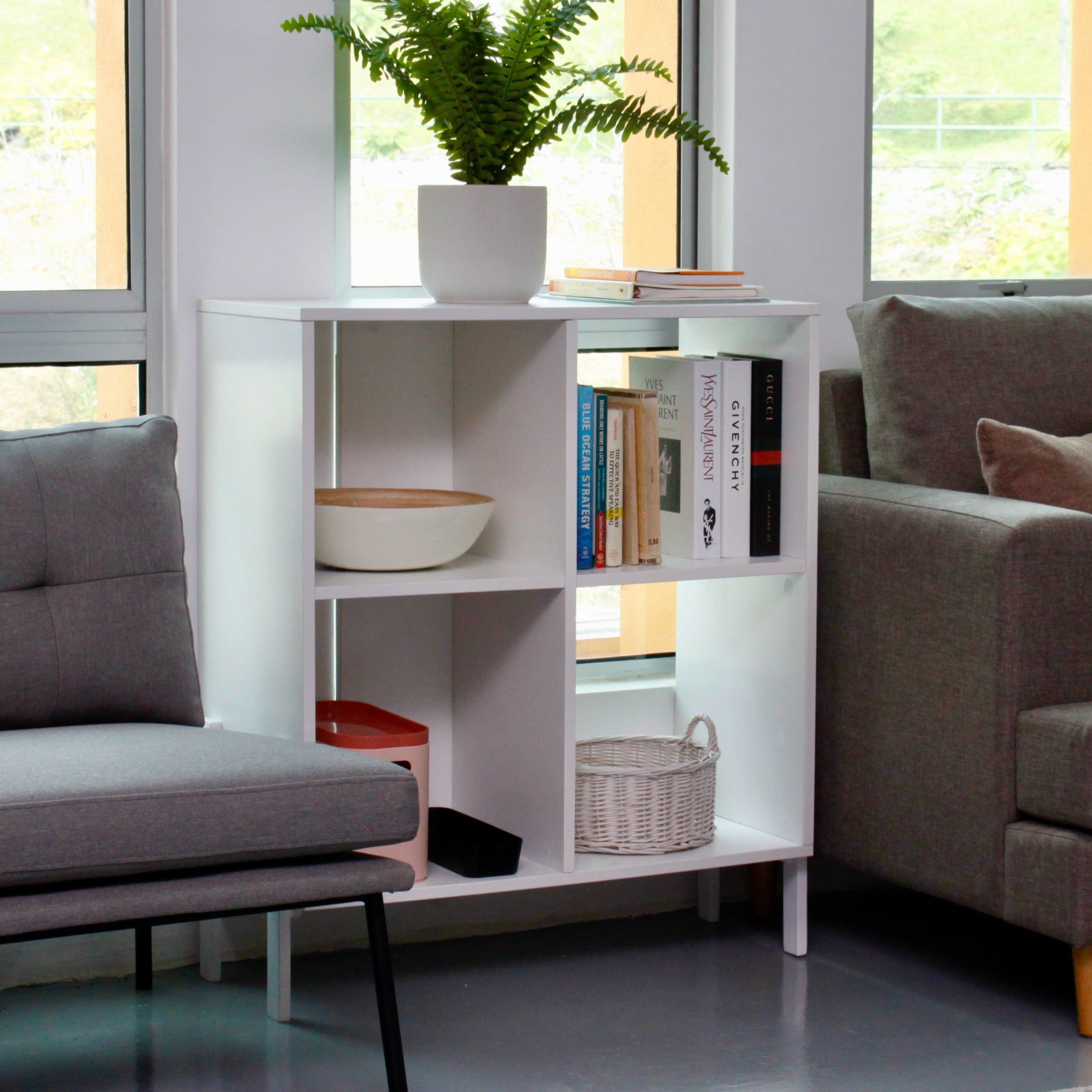 A white CAPI bookcase with four compartments, a fixed shelf, and solid wood legs placed in a living room setting with a plant on top and various items stored.