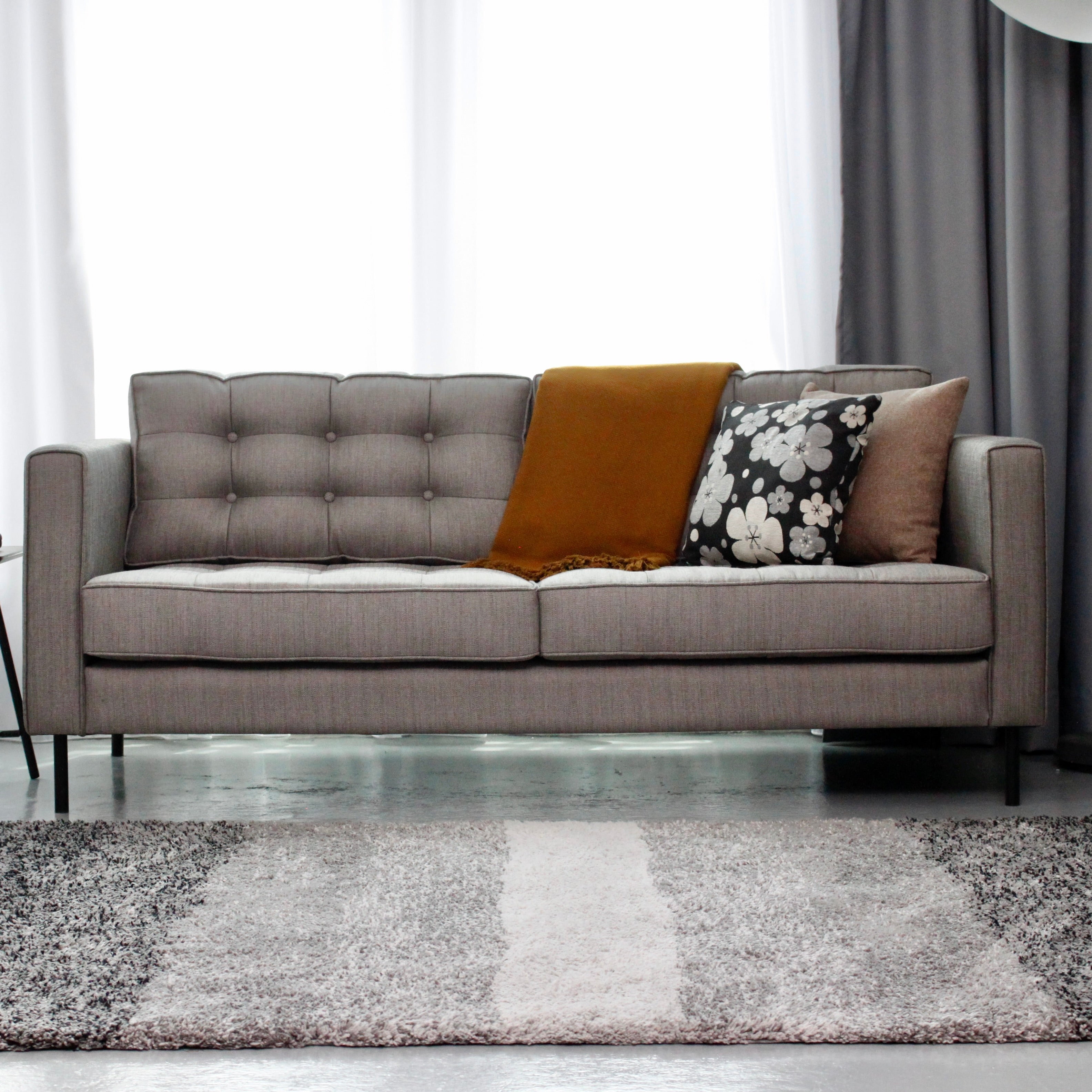 A modern light gray tufted sofa in a styled living room, decorated with a mustard yellow throw pillow, a black and white floral accent pillow, and a brown pillow, set on a striped gray and white shag rug.