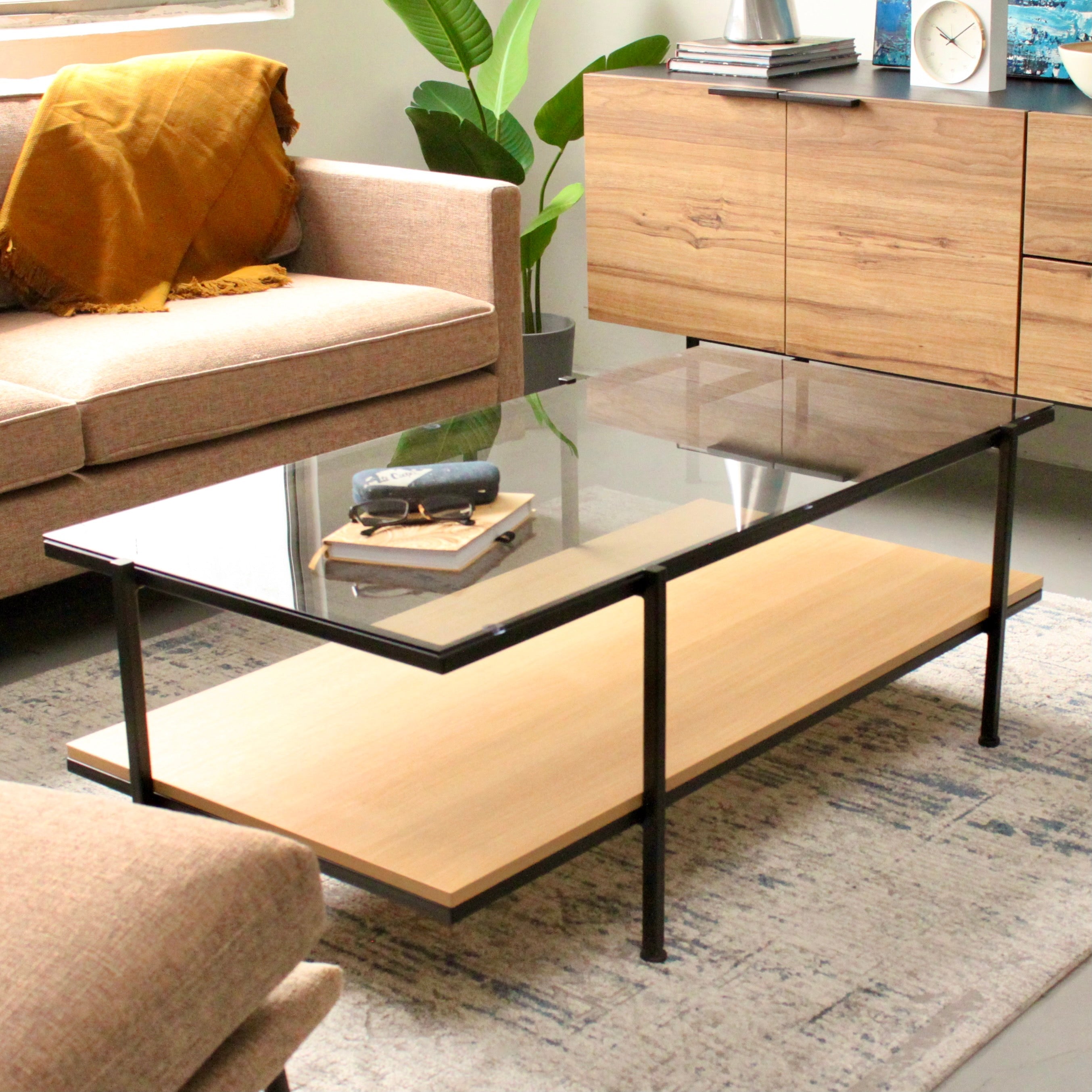 A rectangular coffee table with a glass top and an oak shelf, featuring a black metal frame, placed on a rug in a living room setting with a sofa and plant in the background.