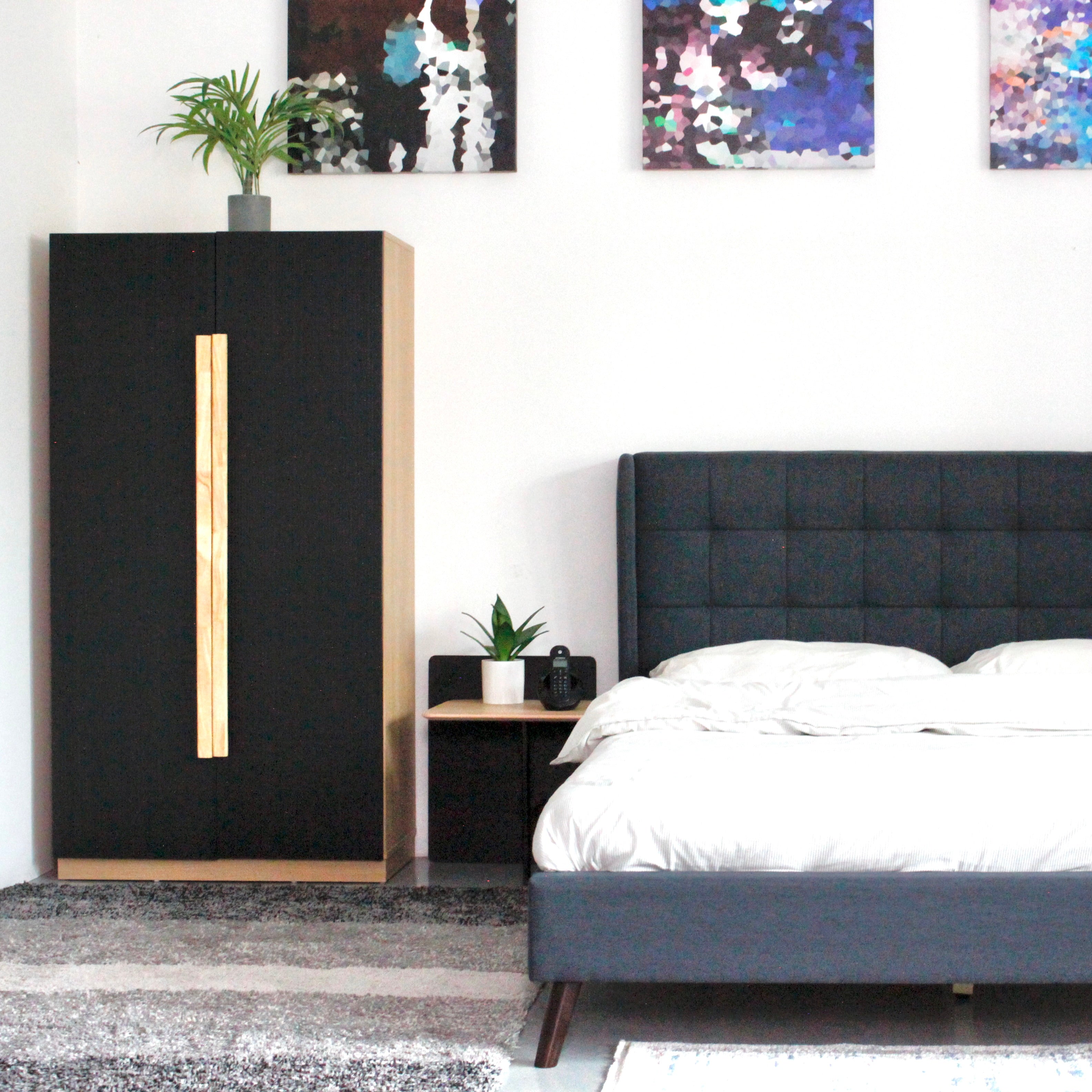 A modern cabinet with black doors and a solid wood handle, placed in a room next to a bed, with a painting on the wall above the cabinet.
