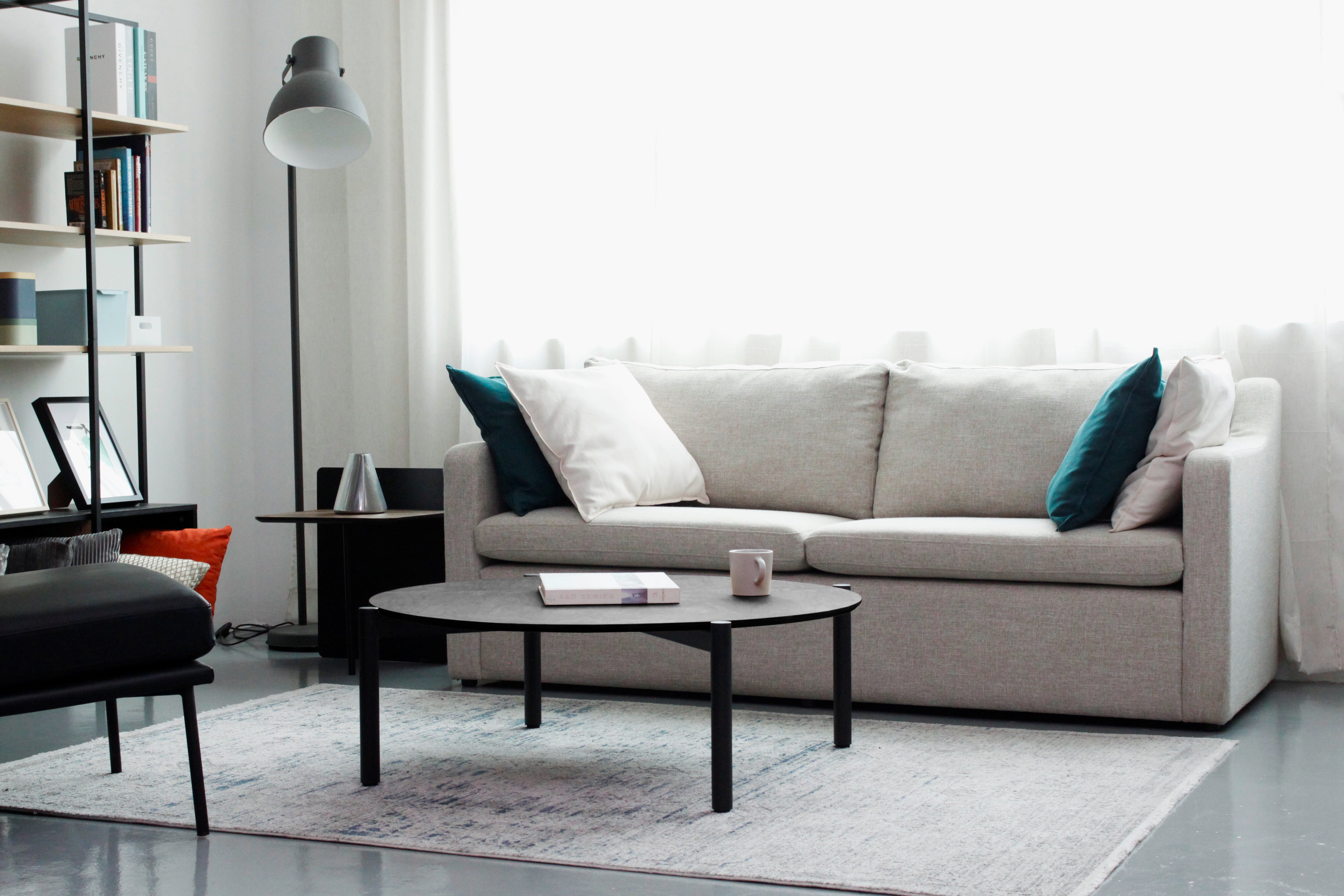 A black round coffee table with a textured top and metal legs, placed on a striped rug in a cozy living room setting with a sofa, armchairs, and decorative items in the background.
