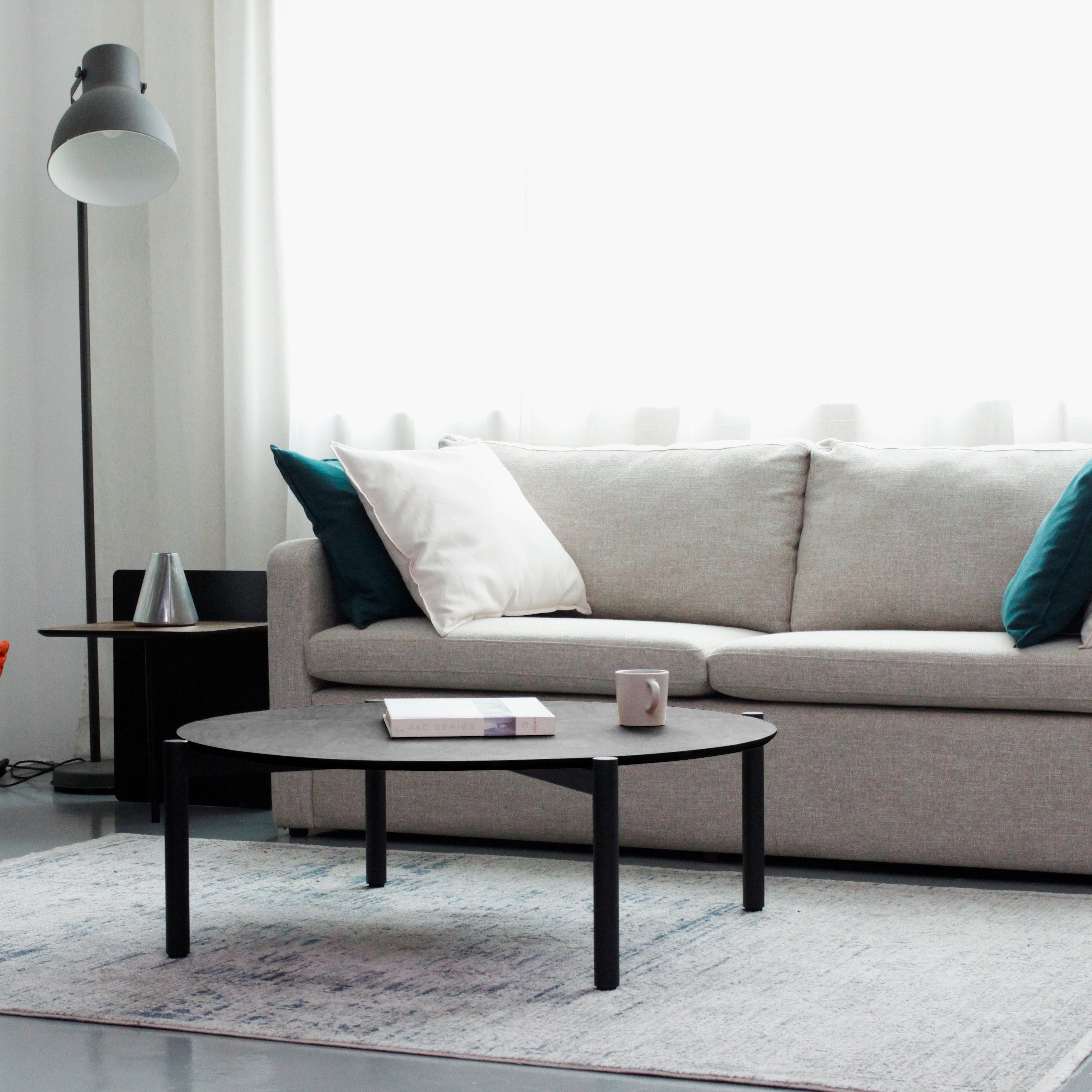 A black round coffee table with a textured top and metal legs, placed on a striped rug in a cozy living room setting with a sofa, armchairs, and decorative items in the background.