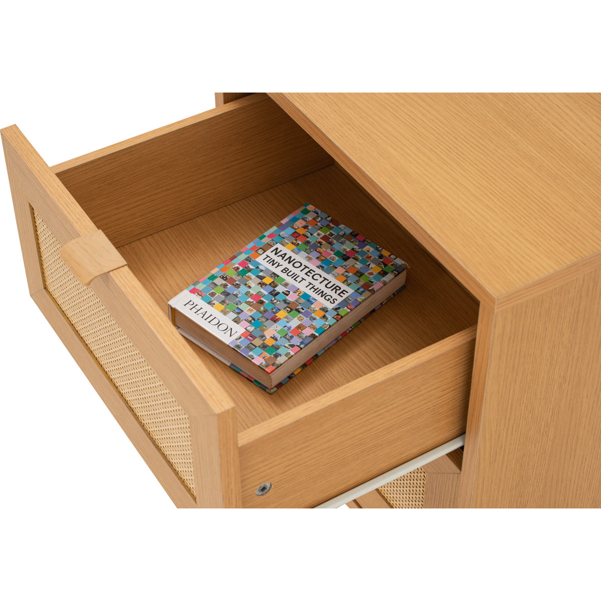 Close-up of a light natural wood drawer with a woven cane pull handle, shown open with a book inside on a white background.