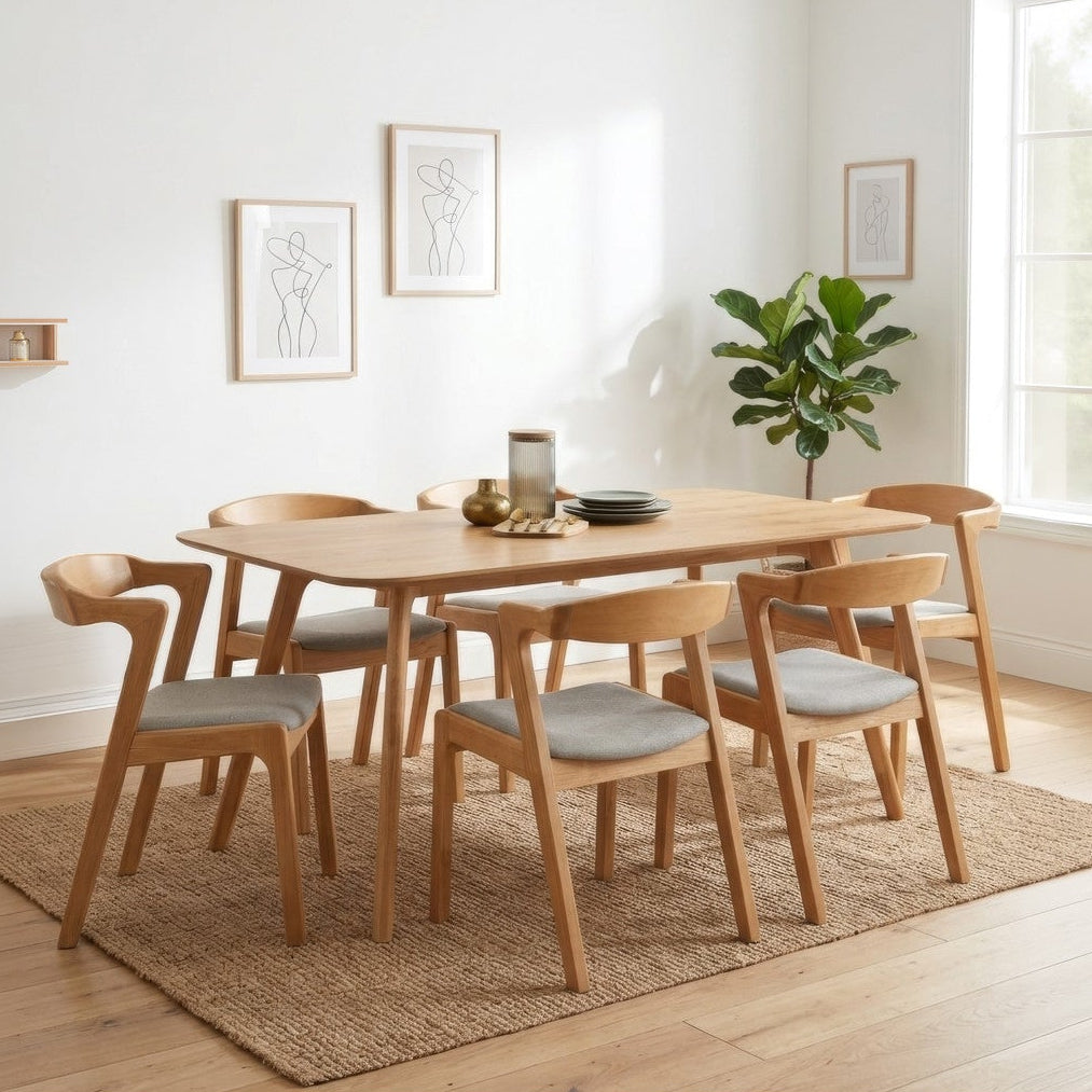 A light oak dining table with six matching wooden chairs featuring light gray upholstered seats, arranged on a natural jute rug in a bright, modern dining room with a window and potted plant.