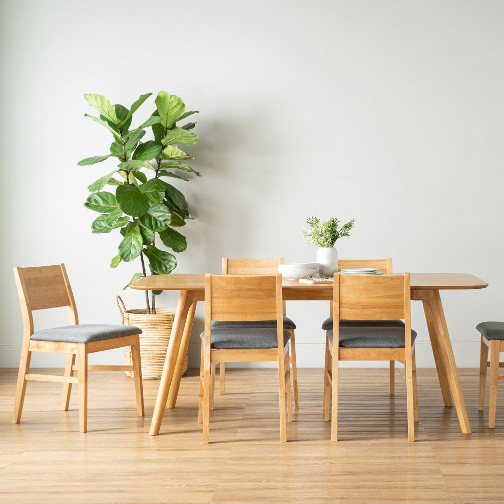 A minimalist dining set consisting of a light oak wood table and four matching chairs with dark gray upholstered seats, arranged in a bright room with a large potted fiddle-leaf fig plant and a small vase of flowers on a light wood floor.