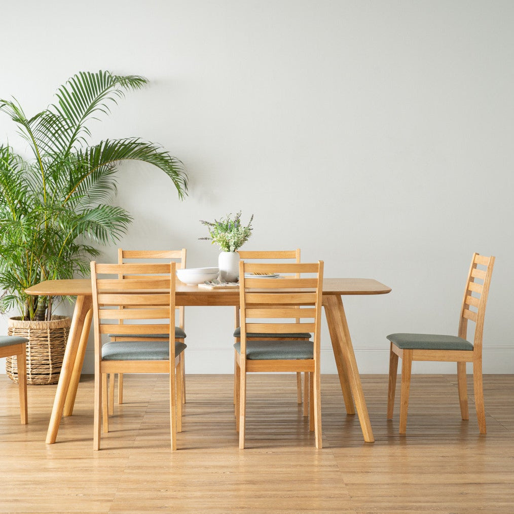 A light natural wood dining set consisting of a rectangular table and four matching slat-back chairs with muted blue-gray upholstered seats, arranged in a minimalist room with a light gray wall, wood floor, and a potted palm plant.