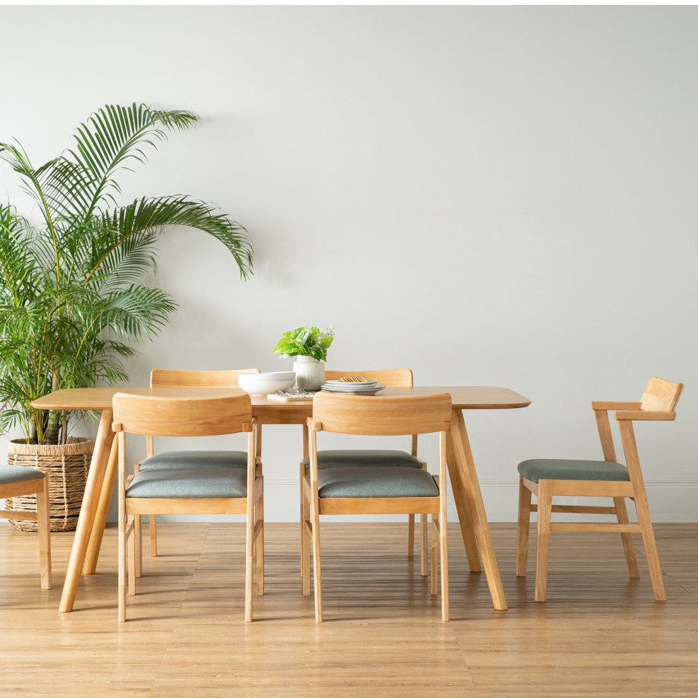 A light oak dining table with four matching chairs featuring gray upholstered seats, arranged in a minimalist room with a light gray wall, light wood floor, and a potted palm plant.
