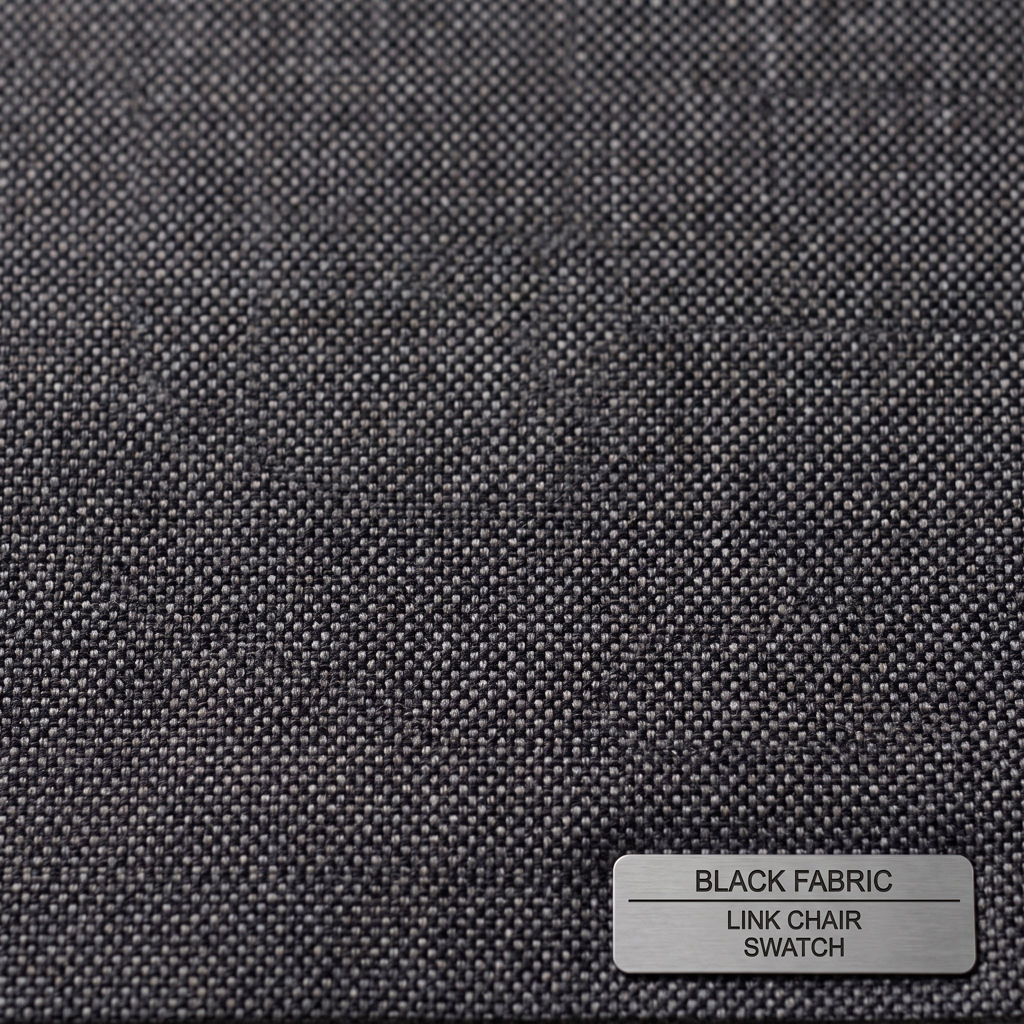 A close-up of a dark charcoal gray woven fabric swatch with a small metal label in the bottom right corner that reads 'BLACK FABRIC LINK CHAIR SWATCH'.