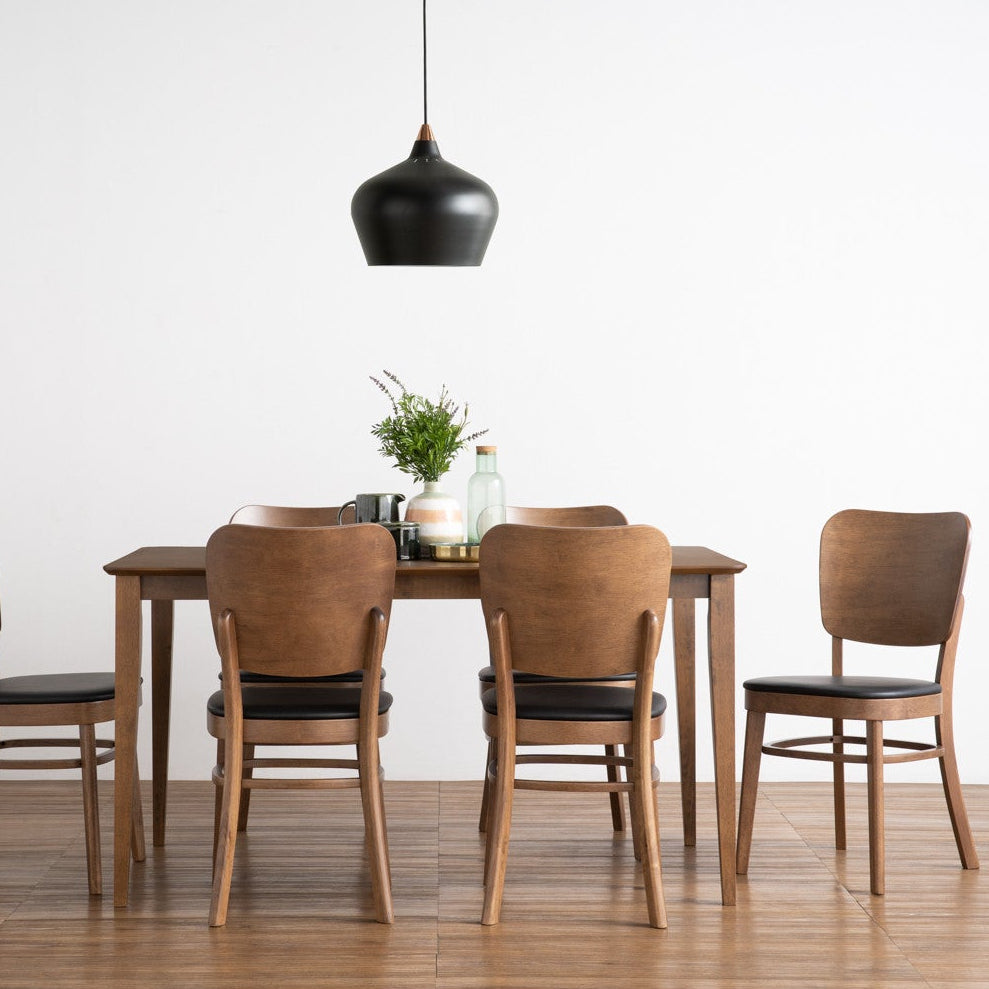 A dining set consisting of a rectangular wooden table and four matching chairs with black seat cushions, arranged in a minimalist room with a white wall and light wood floor under a black pendant light.