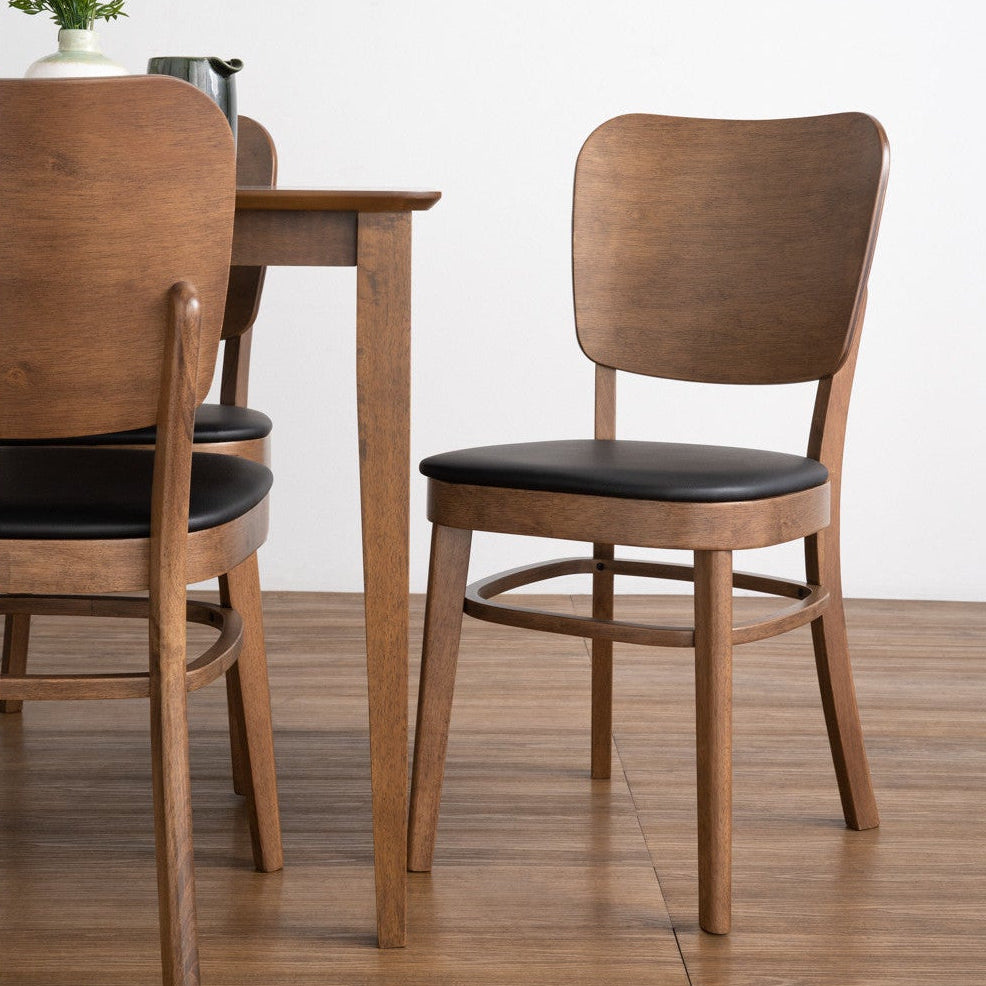 A modern dining room scene featuring a wooden dining table and two matching chairs with black upholstered seats, set against a clean white wall and light wood floor.