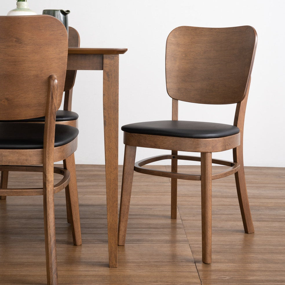 A modern dining room scene featuring a wooden dining table and two matching chairs with black upholstered seats, set against a clean white wall and light wood floor.