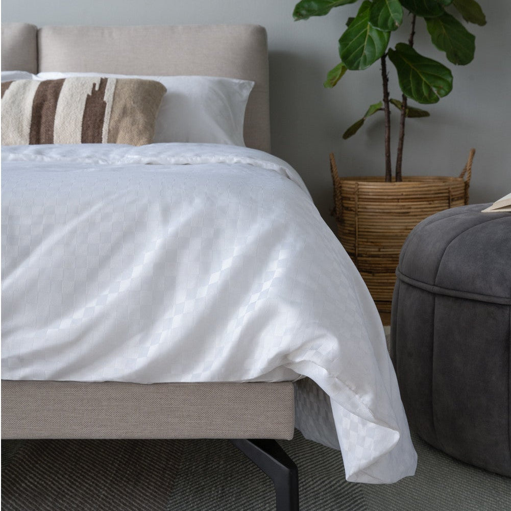 A modern bedroom scene featuring a bed with white bedding and a textured beige upholstered frame, styled with a striped accent pillow, next to a gray ottoman, a potted fiddle-leaf fig plant in a wicker basket, and a gray patterned rug.