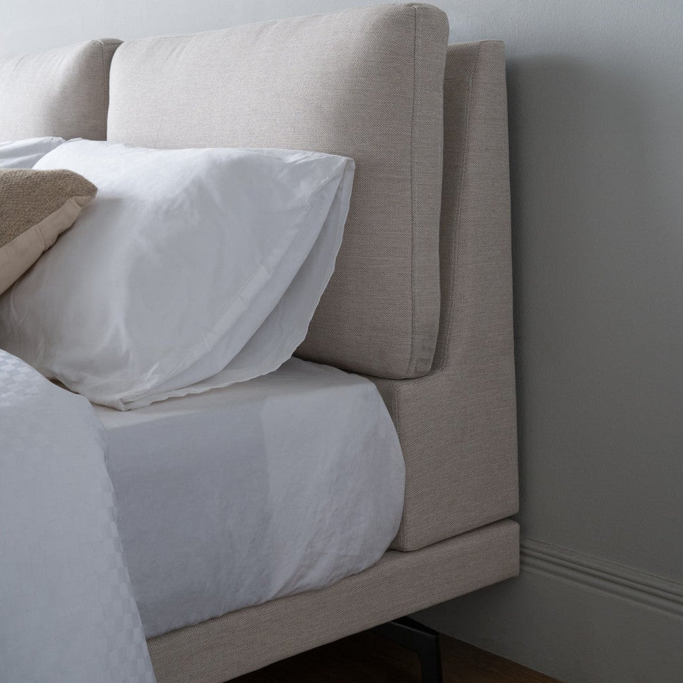 A close-up of a modern upholstered bed headboard in a light beige fabric, styled with white bedding and pillows against a neutral gray wall.