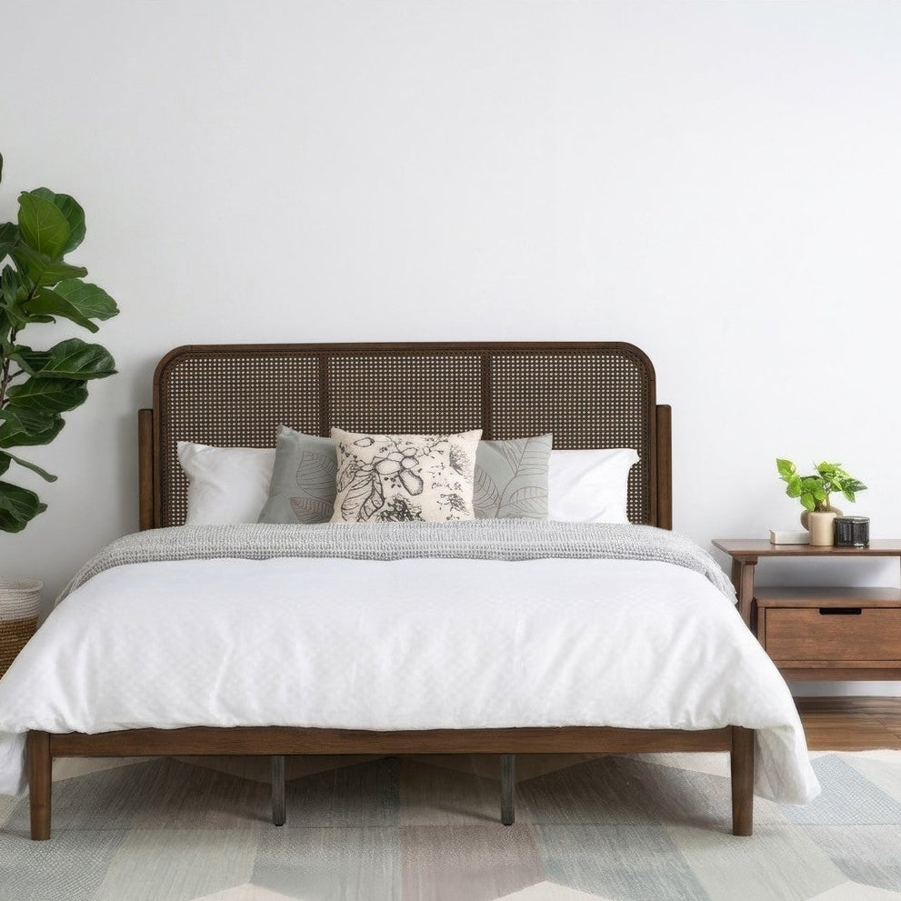 A mid-century modern wooden bed frame with a woven cane headboard, styled with white bedding and decorative pillows in a minimalist bedroom setting with a plant and nightstand.