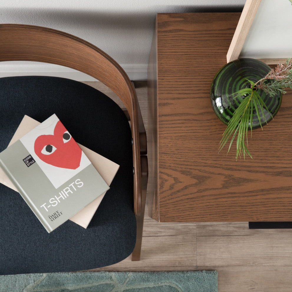 Close-up of a wooden chair with a magazine and a wooden side table with a plant on a light wood floor.