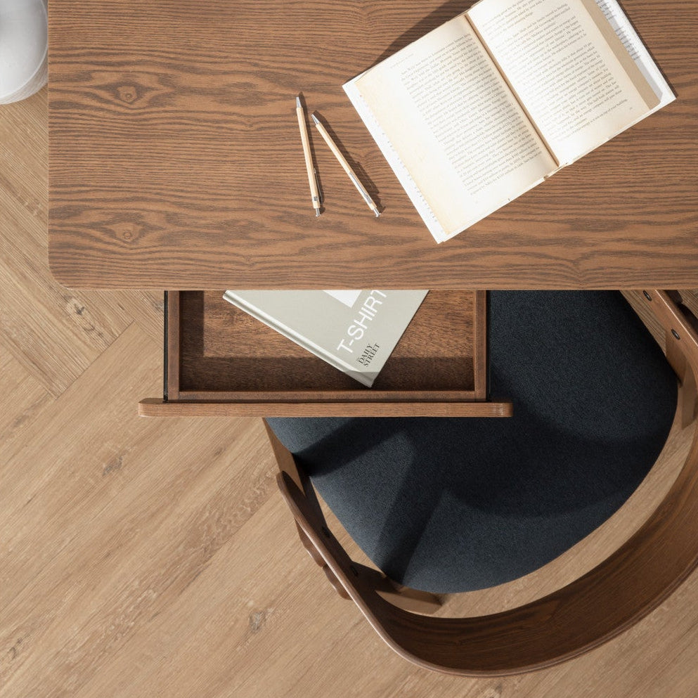 Wooden desk with an open book, pen, and small plant on a light wooden floor.
