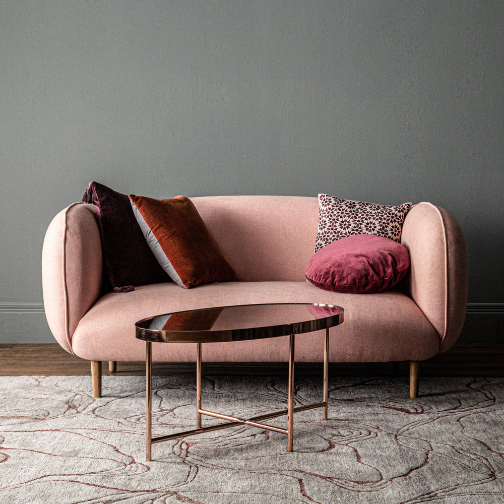 Pink sofa with decorative pillows and a small round table in a room with a gray wall and patterned rug.