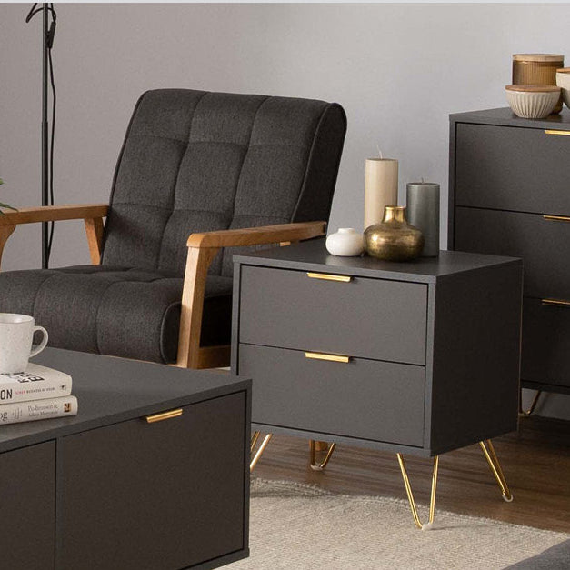 A dark gray side table with gold metal legs and gold drawer pulls, shown in a styled living room setting next to an armchair and a larger matching cabinet. The image has a text overlay at the bottom reading 'VOLOS Side Table'.