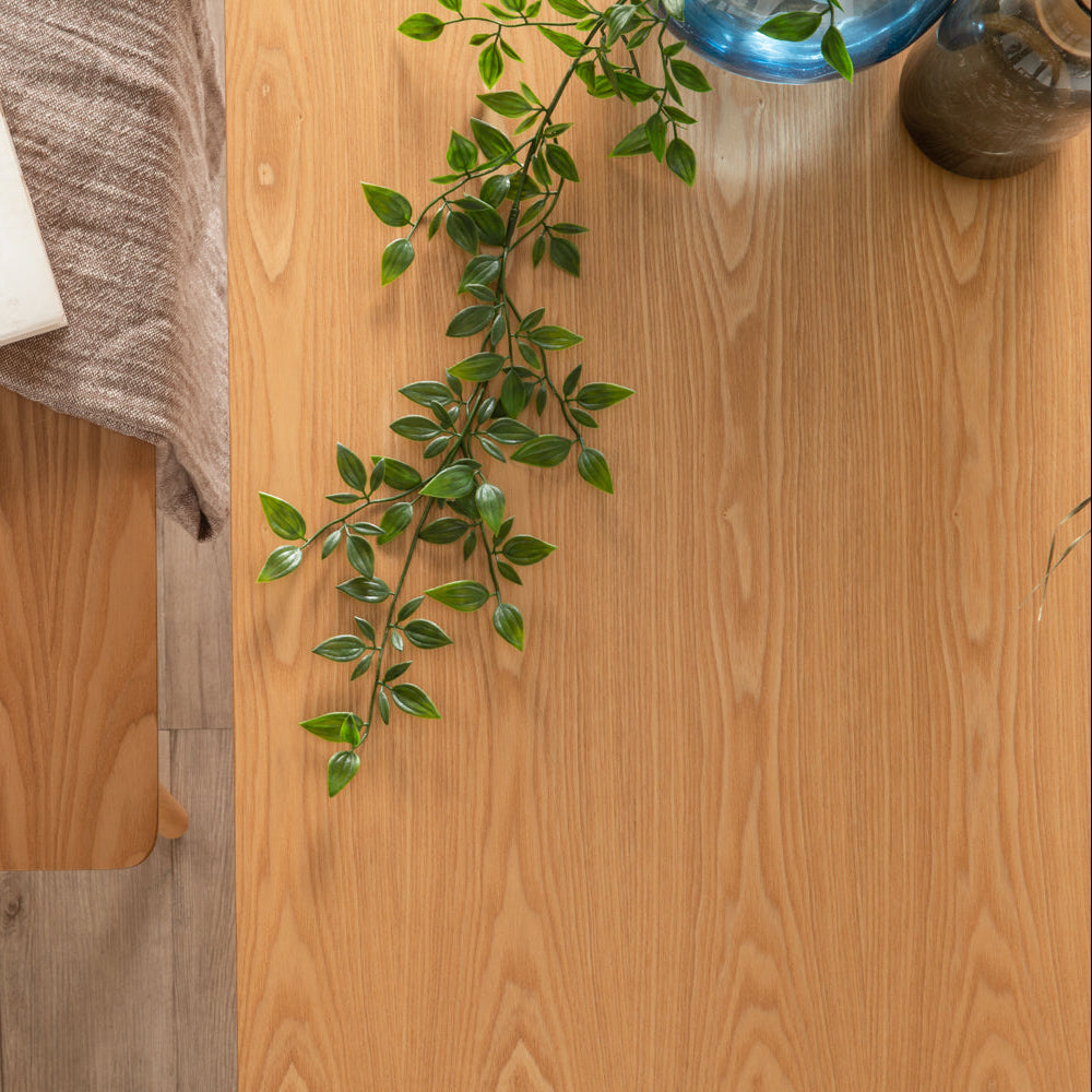 A close-up of a light oak wood grain surface, likely a tabletop or flooring sample, styled with a green leafy vine and partial home decor props.