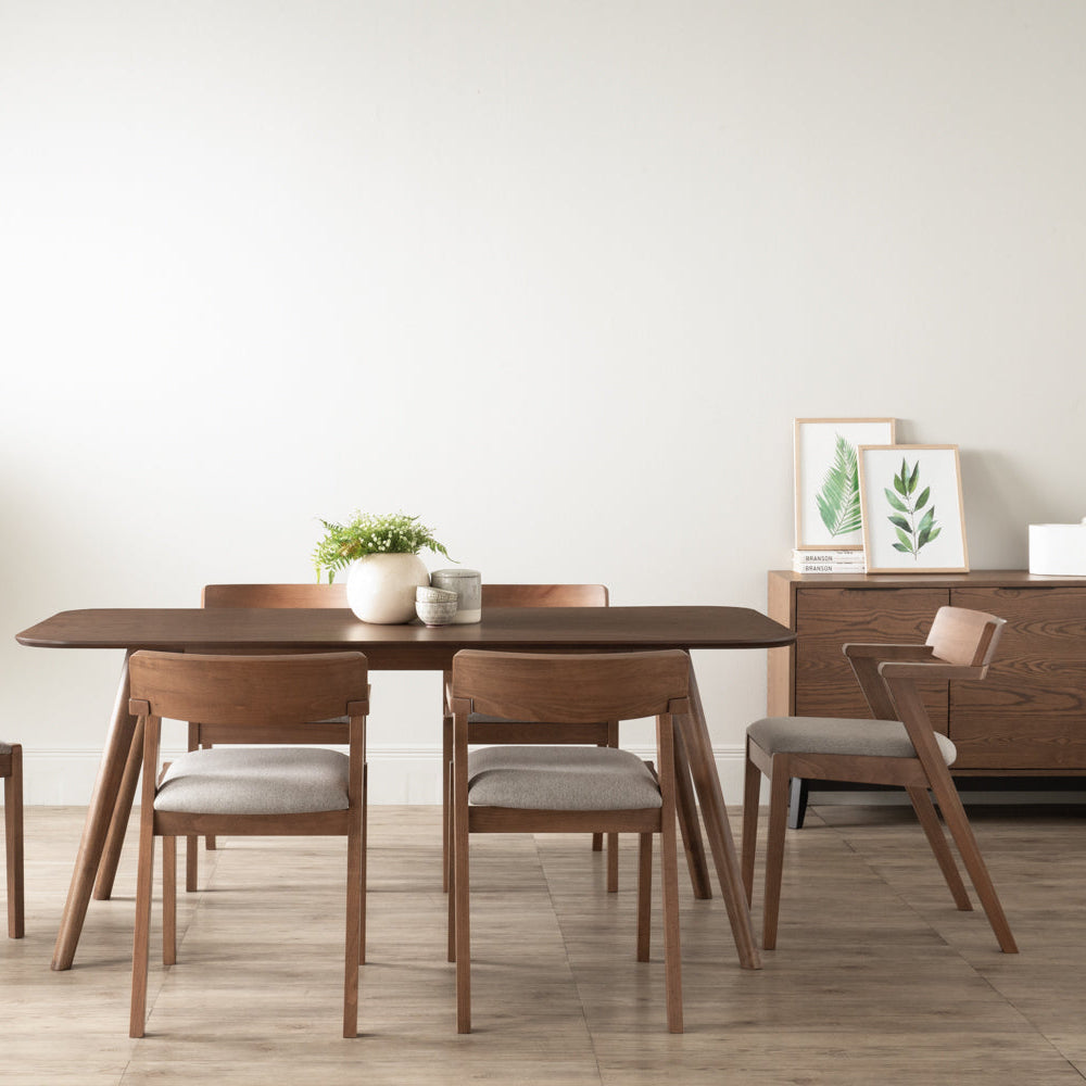A mid-century modern wooden dining table with four matching chairs featuring light beige upholstered seats, set in a minimalist dining room with a white wall and light wood floor.