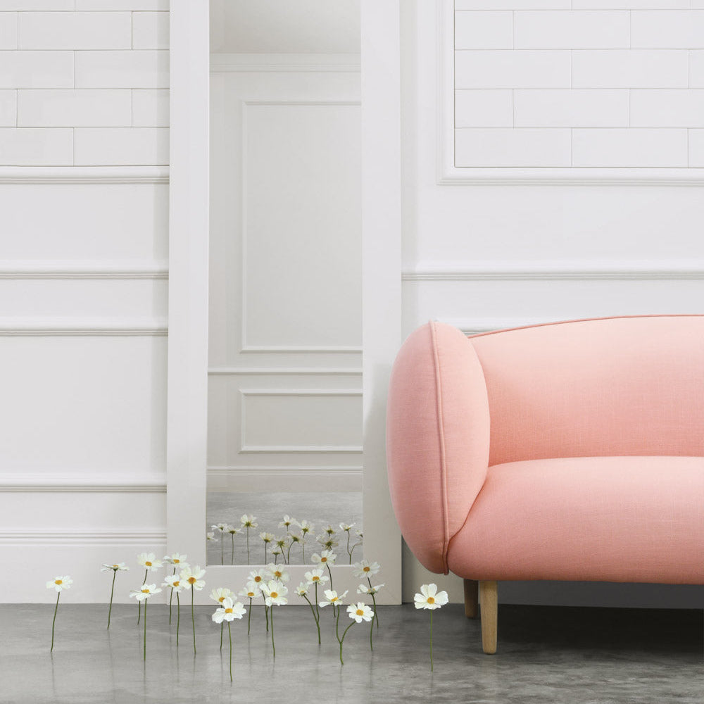 Pink sofa against a white paneled wall with a row of white flowers on a gray floor.