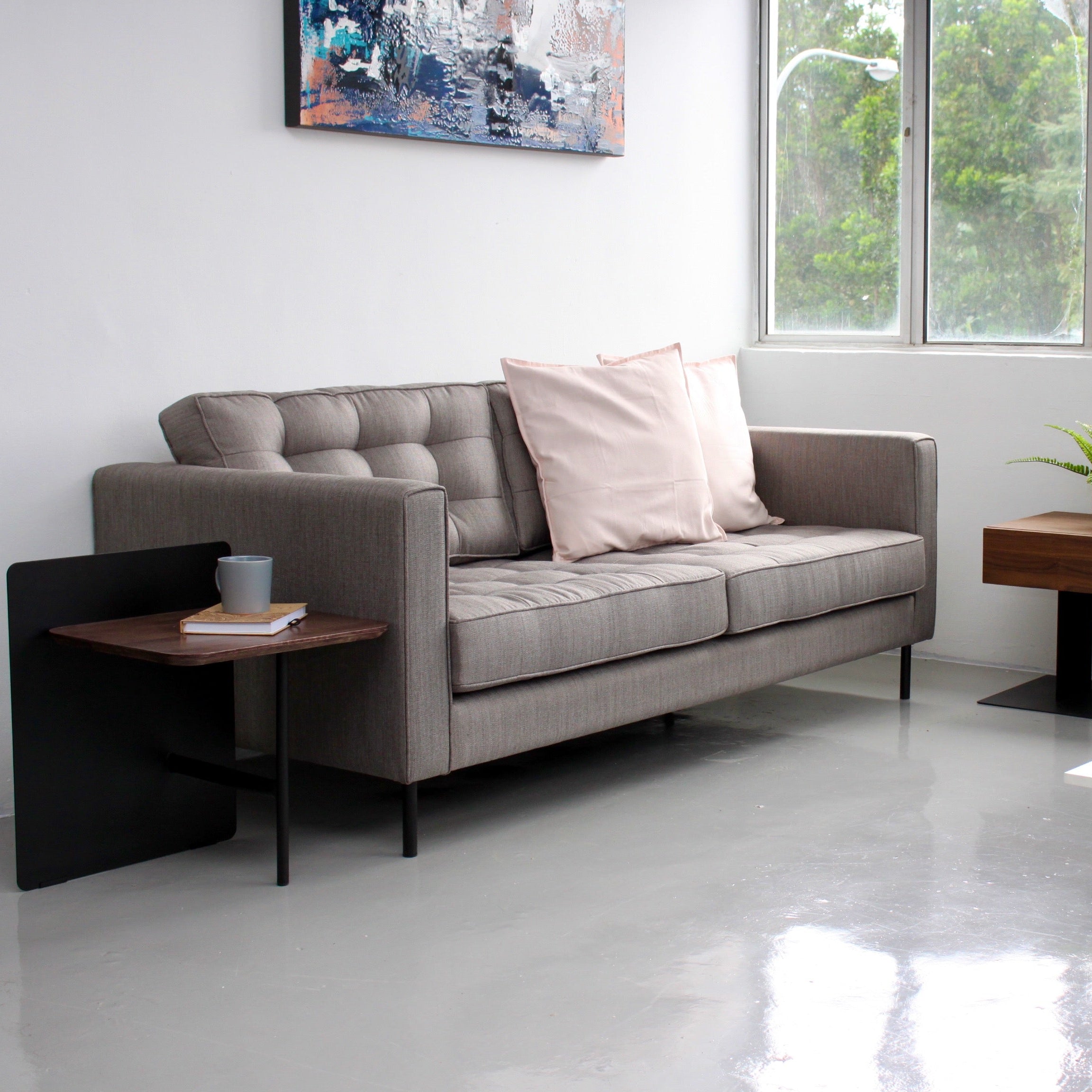 A modern gray tufted sofa in a bright, minimalist living room setting with a light pink pillow, flanked by black and wood side tables.