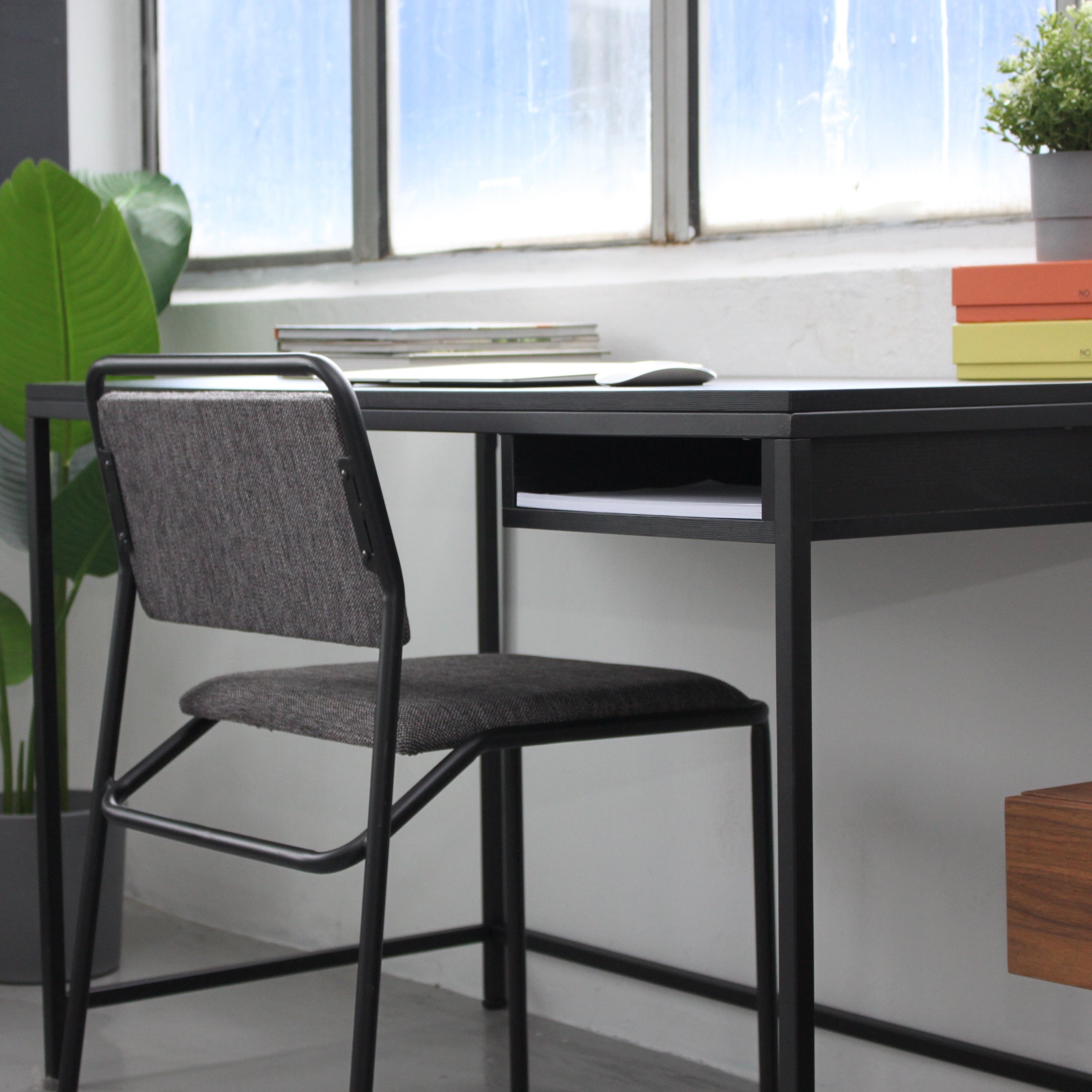 A modern all-black desk with a drawer and metal legs, accompanied by a mesh chair, set against a window in an office environment.