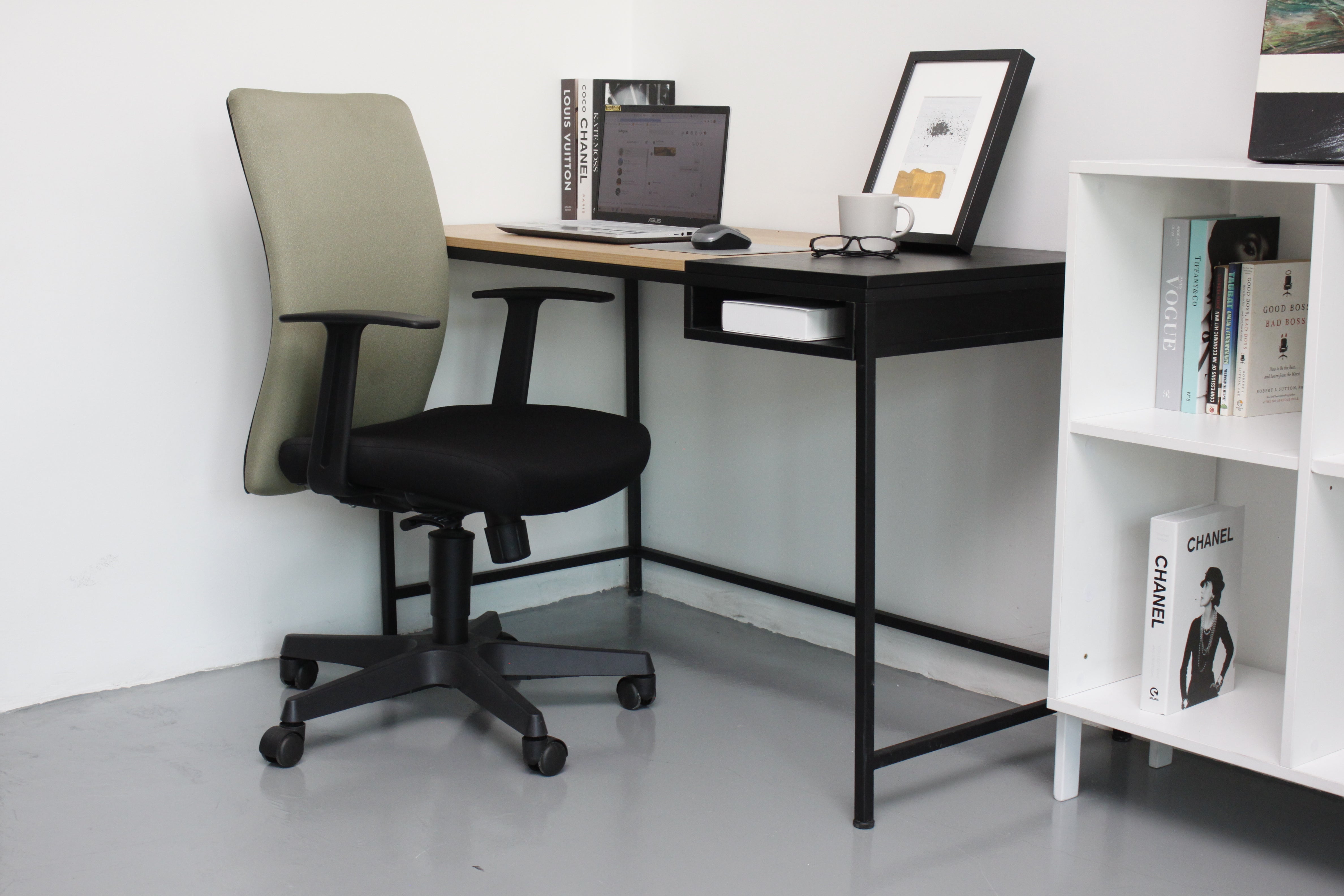 A black office chair with a green fabric backrest, a metal five-pronged base, and a rectangular desk with a laptop, books, and a framed picture in the background.