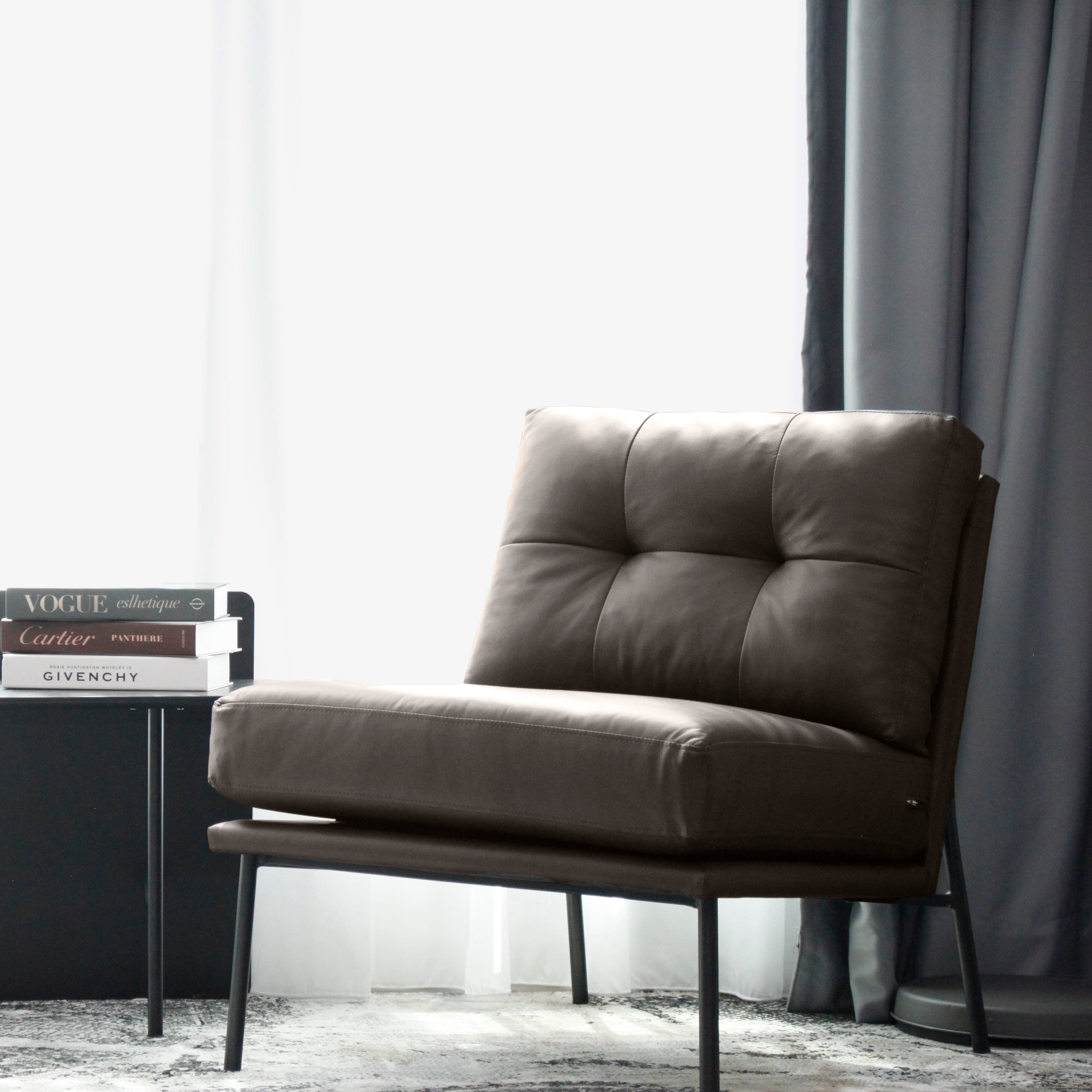 Modern taupe armchair in a room with a curtain and a black side table.