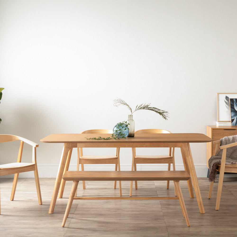 A light natural wood dining table set in a minimalist room, accompanied by a matching wooden bench and chairs. The table is styled with a small vase of greenery, set against a clean white wall and light wood flooring.