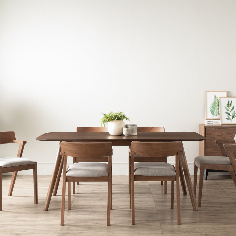 A mid-century modern wooden dining table with four matching chairs featuring light beige upholstered seats, set in a minimalist dining room with a white wall and light wood floor.