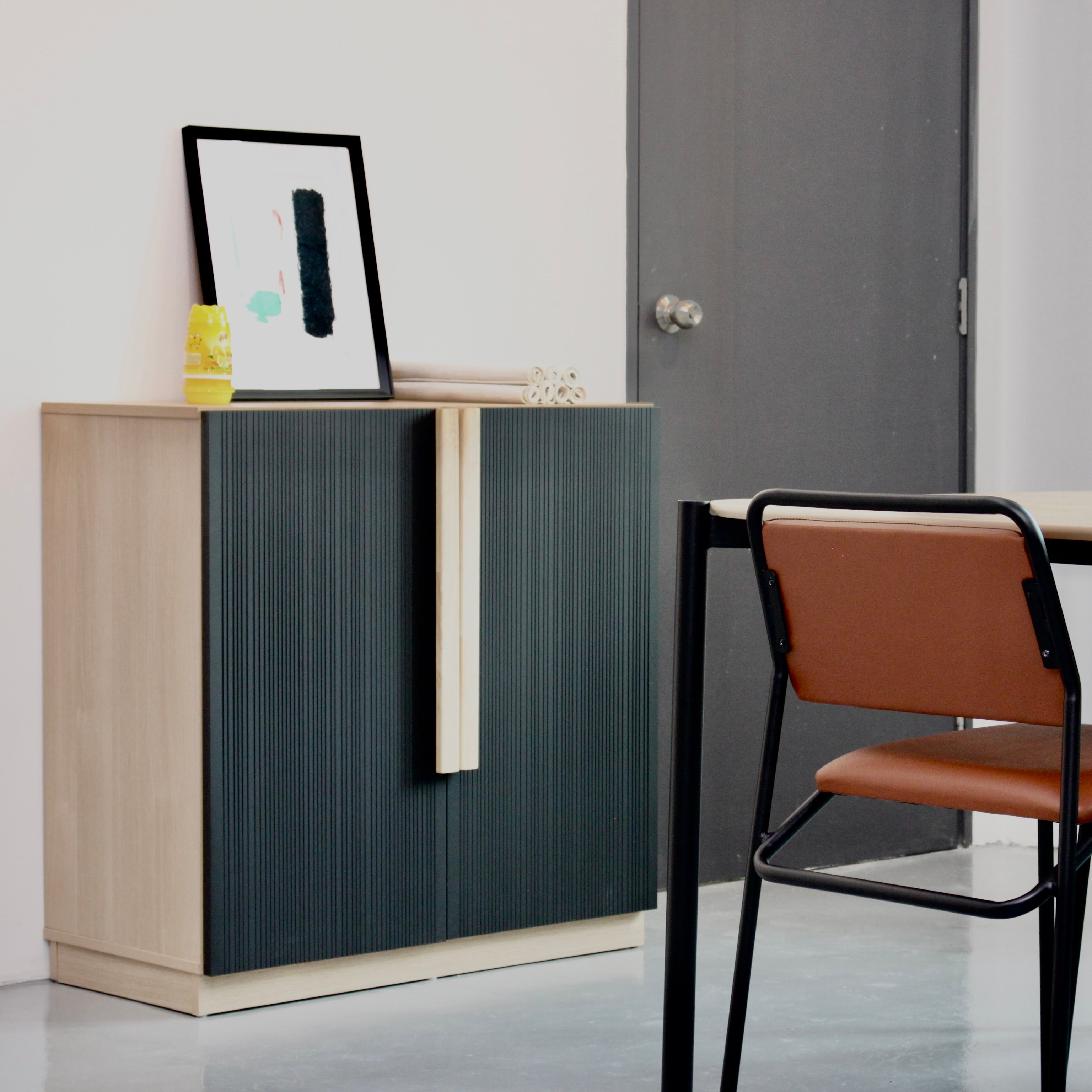 A cabinet with black doors and a natural oak wood handle, positioned against a wall with a chair in front of it. The cabinet has a picture frame on top and a photograph of a plant on the handle.